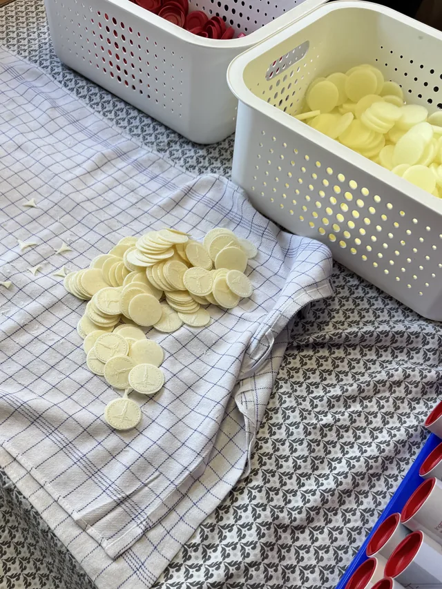Pale round communion wafers piled on a checked cloth beside a basket of wafers and packaging containers.