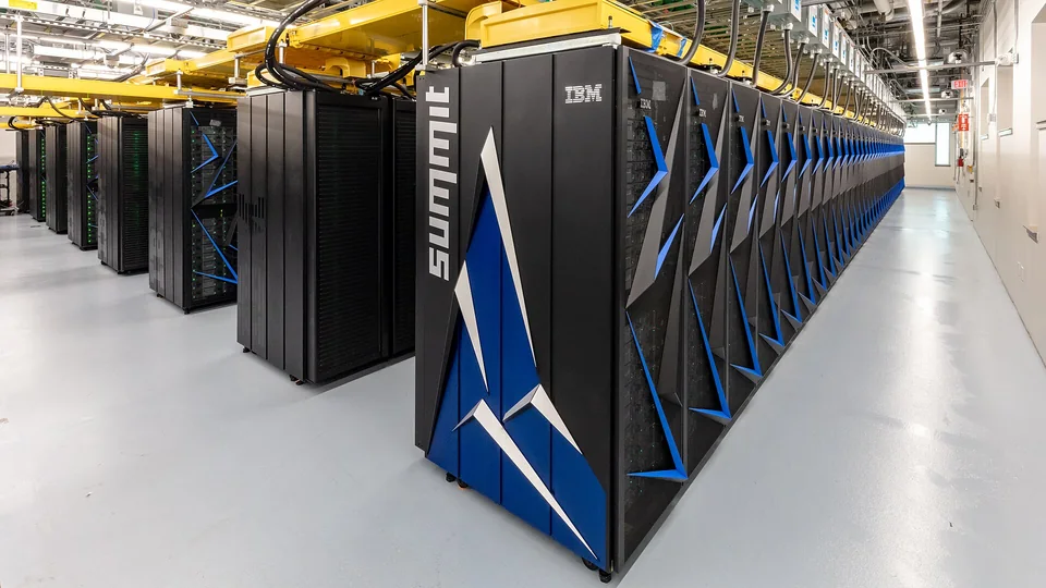 Rows of black and blue Summit supercomputer cabinets recede across a brightly lit machine room.