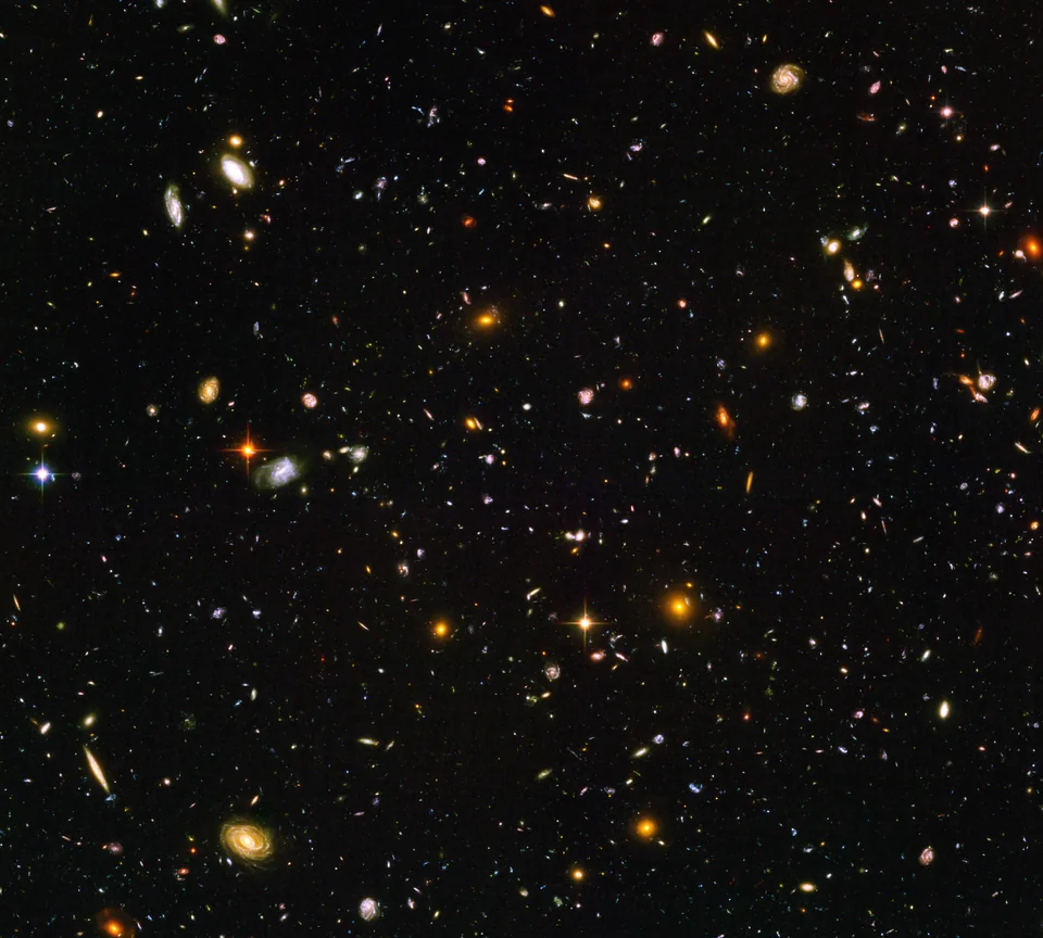 A black field filled with hundreds of tiny red, blue, and golden galaxies, with a few larger spirals.