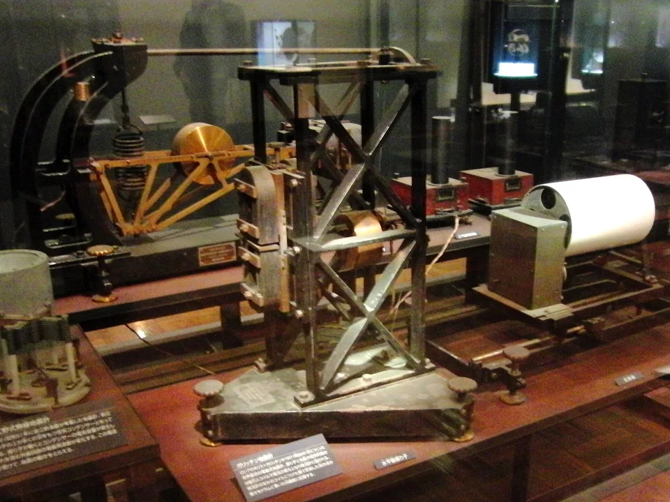 A preserved Galitzin seismograph with suspended mechanical components, a dark metal frame, and recording equipment on a museum display base.