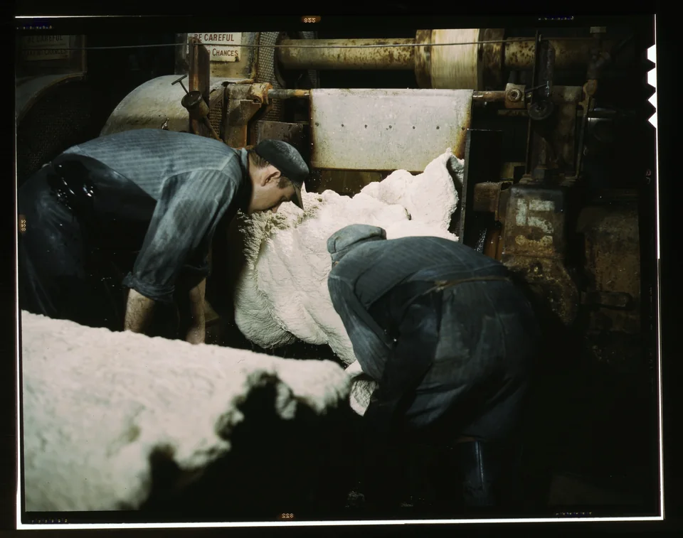 Two workers guide a pale sheet of synthetic rubber from a rolling mill in a dark factory.