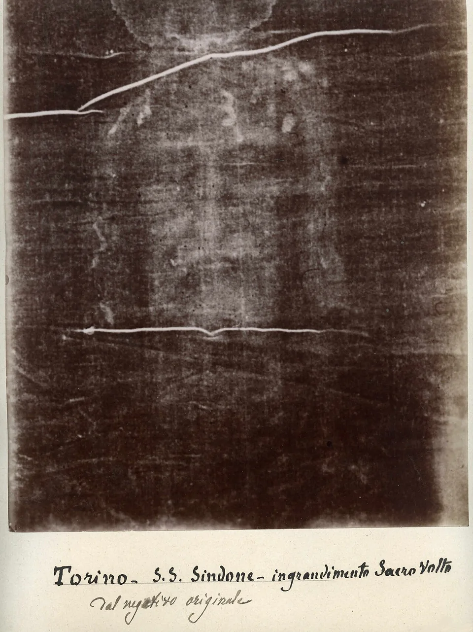 A sepia photographic negative of the face on the Shroud of Turin, crossed by visible folds in the cloth, with a historical handwritten caption below.