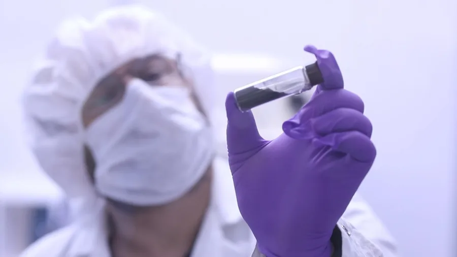 Jason Dworkin, wearing a mask and cleanroom clothing, holds a clear vial of dark Bennu material between purple-gloved fingers.