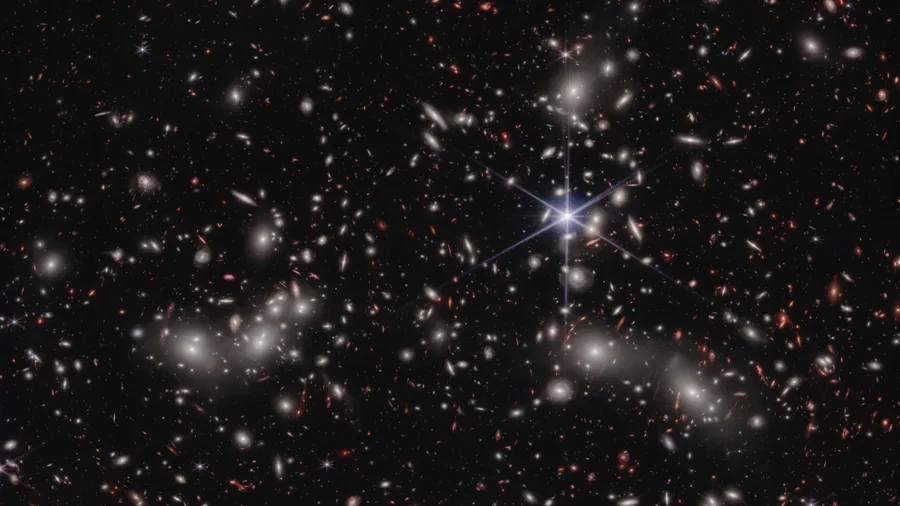 A dense field of white, red, and gold galaxies against black space, with a blue diffraction-spiked foreground star right of center.