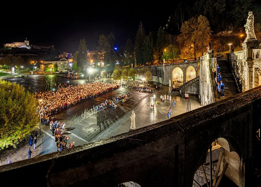 Pilgrims gather at the illuminated sanctuary in Lourdes beneath a blue evening sky.