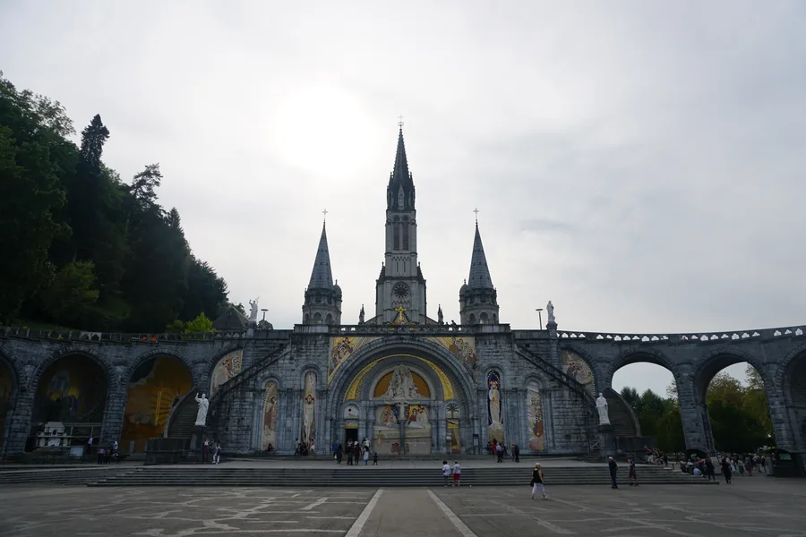 An open plaza faces the Rosary Basilica’s decorated façade and sweeping stone arcades, with tall church spires rising against an overcast sky.