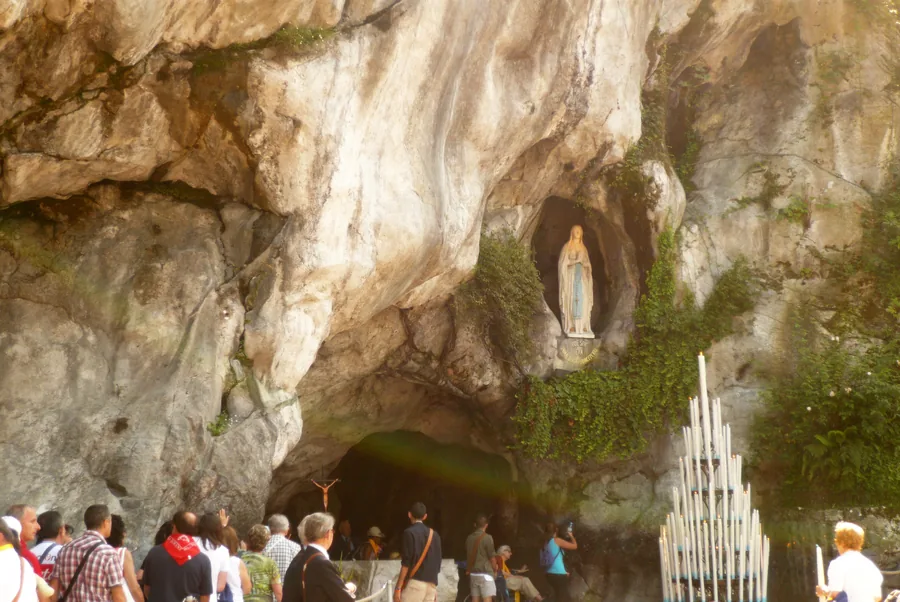 Visitors gather below the rocky Massabielle grotto, with a Marian statue in a niche and a tall stand of candles at right.