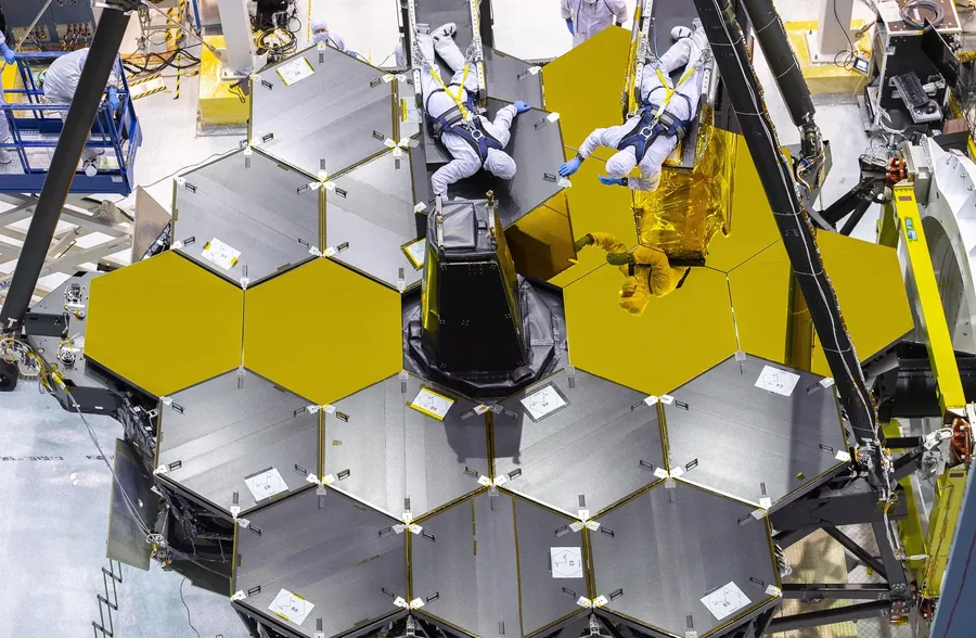 Two technicians in cleanroom suits remove covers from Webb's hexagonal mirrors, revealing gold surfaces beneath.