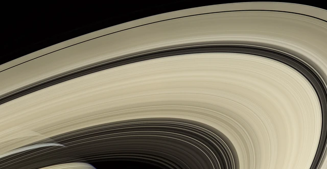 Wide, cream-colored arcs of Saturn’s rings curve across black space, with darker gaps and faint clouds visible through the inner rings.